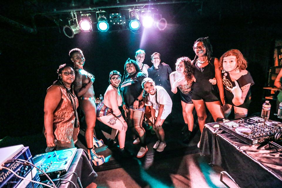 A group of people dressed for a dance party and standing behind tables of DJ gear.