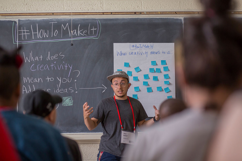 View from behind blurred focus attendees of an AMC session, with a presenter in front with a chalkboard with questions about creativity