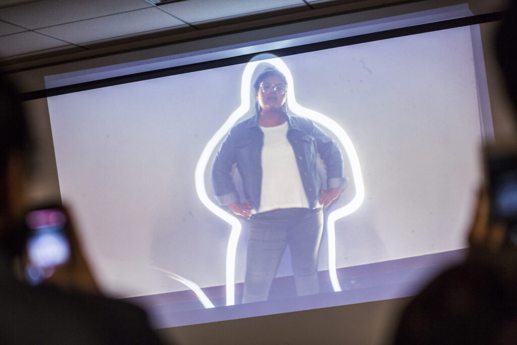 Image of a standing person with arms akimbo of color projected onto a screen outlined by a streak of light