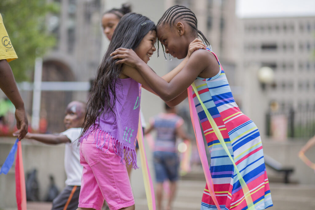 Two girls of color playing outdoors at the AMC, smiling with their foreheads touching and hands on each others shoulders, wearing bright colored clothing