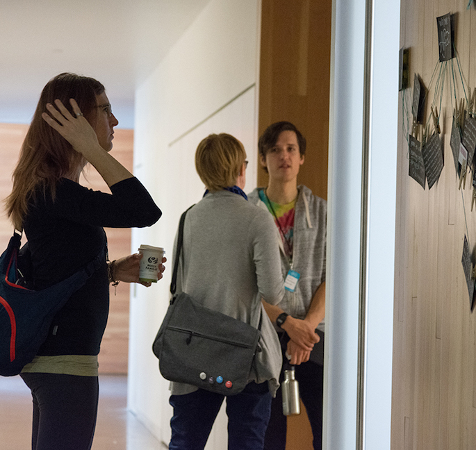 Two people in a conversation at the Affect Conf with a third person reflecting on writing pinned to the wall