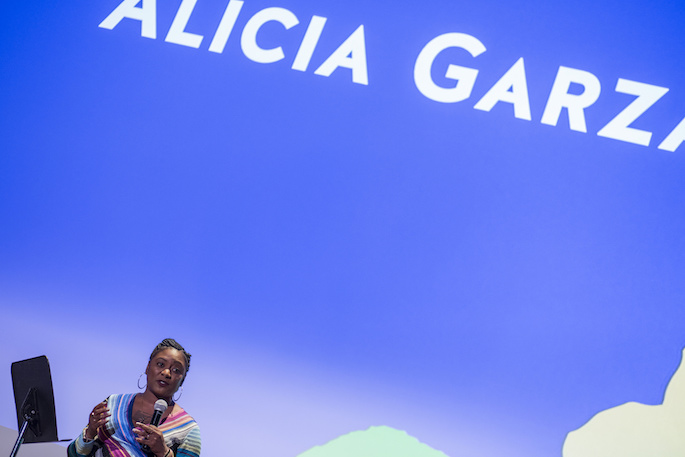 Alicia Garza speaking alone on stage in front of a blue backdrop at the opening ceremony of the 2017 AMC