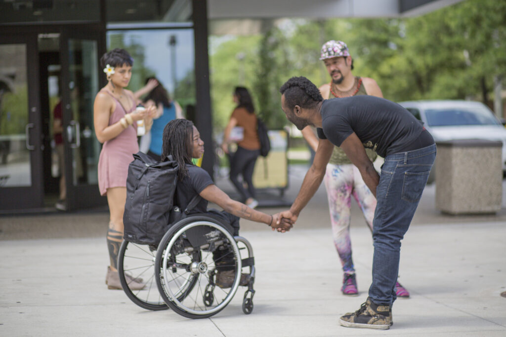 Four people meeting each other outside at the AMC, one in a wheelchair shaking hands with another standing