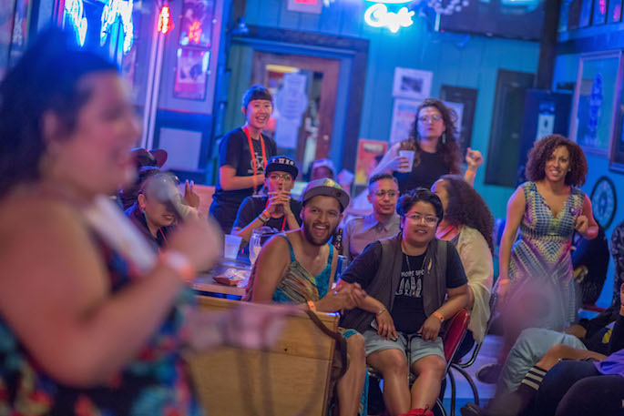 Close up of a person singing karaoke in front a neon sign lit bar with a small group cheering