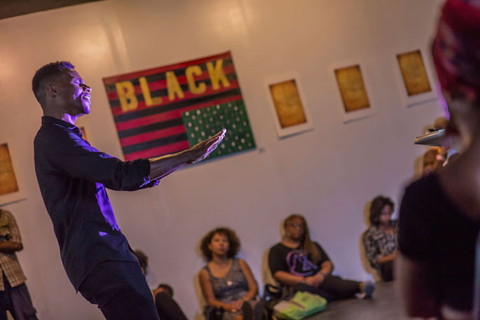 A solo performer singing in a gallery with people sitting on the floor along the wall watching