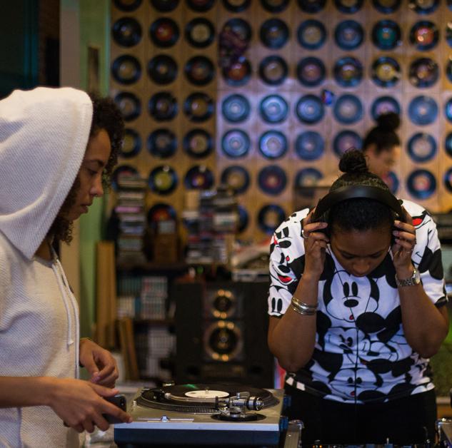 Two Black DJs at a table with a wall of records in the background