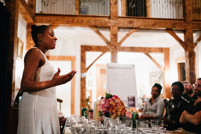 Cornetta standing at a table with fancy drinking glasses and flowers, speaking with hand guestures in front of a small group of people