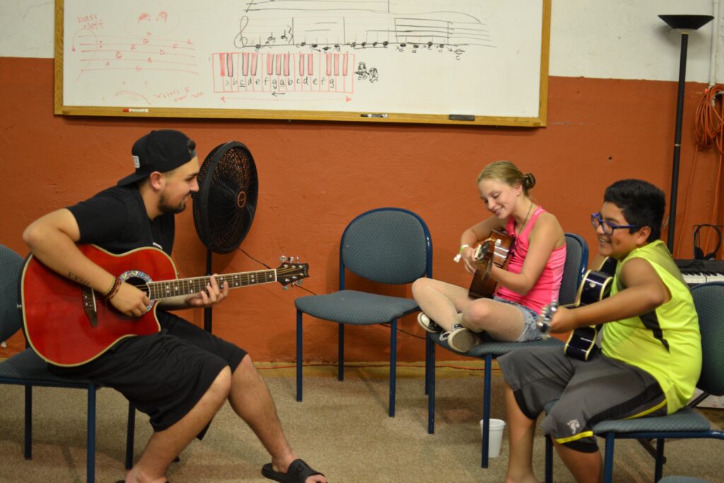 A young Latinx man teaching two younger people how to play guitar, each of them holding their own guitar