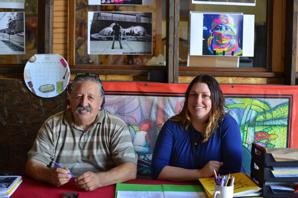Founders of Garage Cultural Ismael and Amelia Duran seated at a desk in front of a wall with various pieces of art and photography