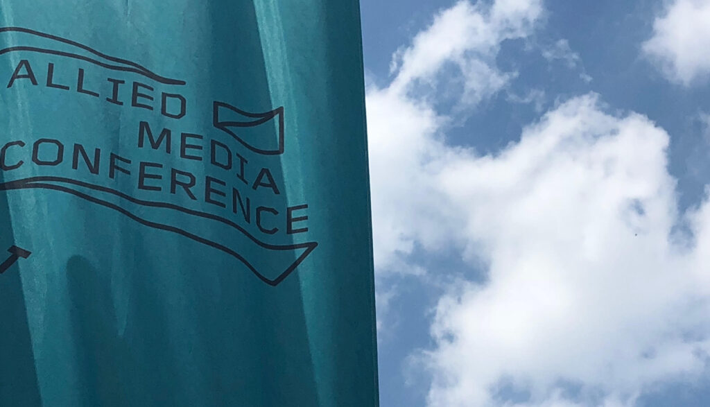 Teal Allied Media Conference flag with white clouds and a dark blue sky