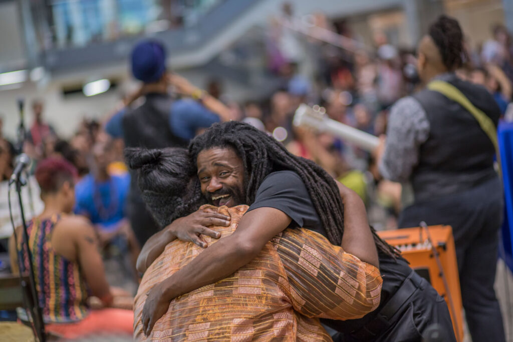 Two Black attendees of the AMC hugging and smiling in the foreground with a band facing the other way behind them and blurry