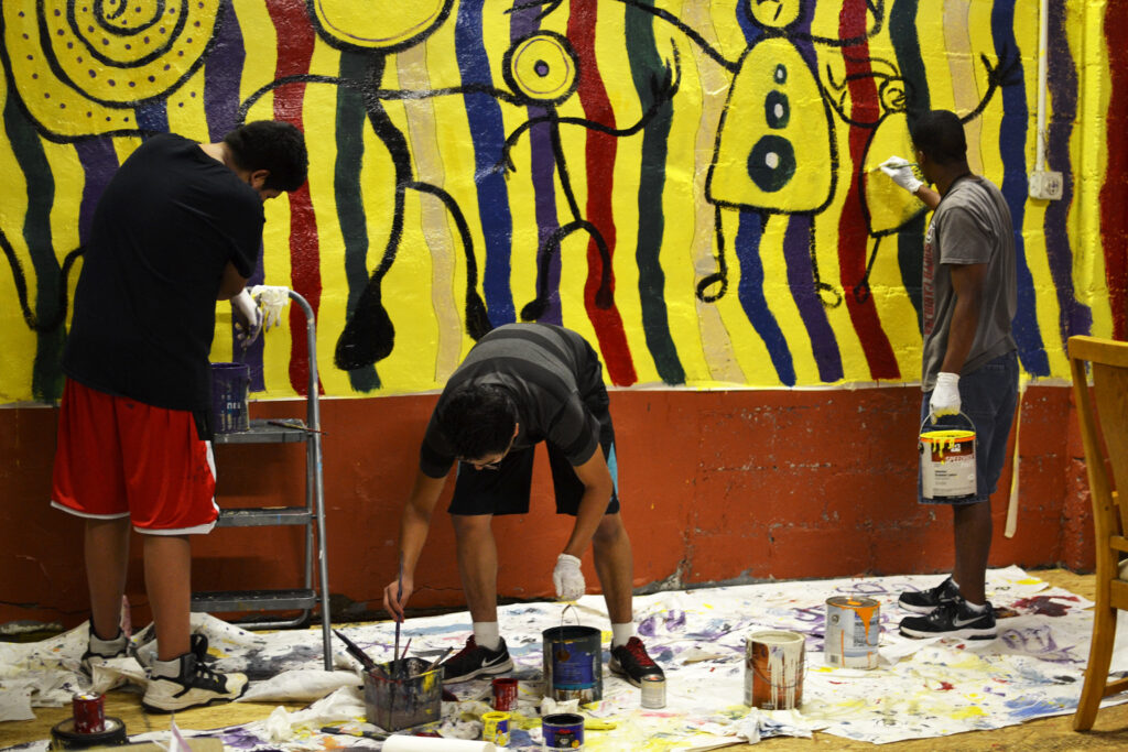 Three young men of color painting a mural indoors of colored vertical stripes and abstract people