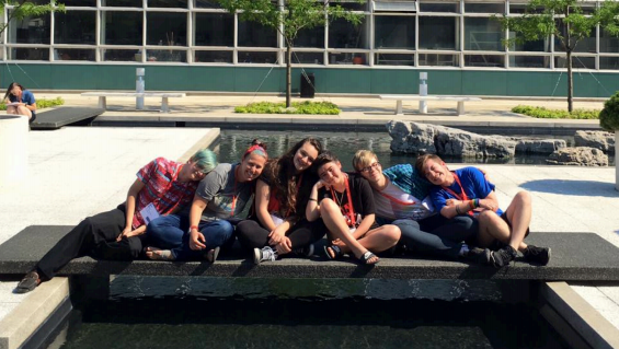 Members of the Bloomington Queer Youth Delegation outside of the AMC posing together sitting cross legged and leaning in on each other in a row