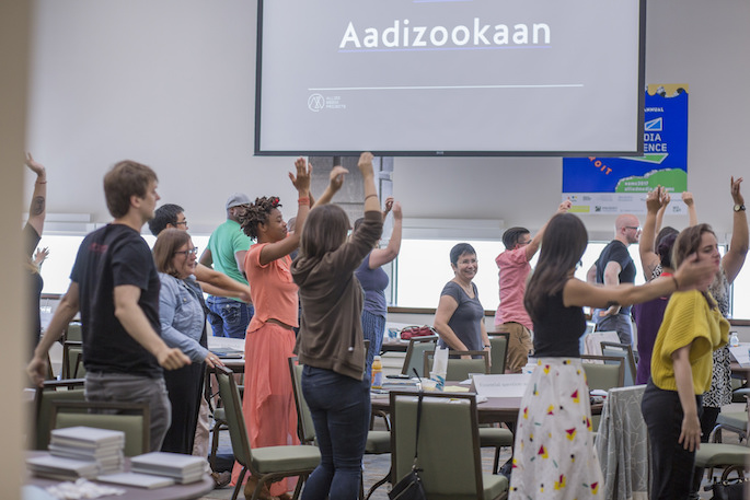 AMC attendees in a classroom space in an activity with their hands in the air and "Aadizookaan" on the screen behind them