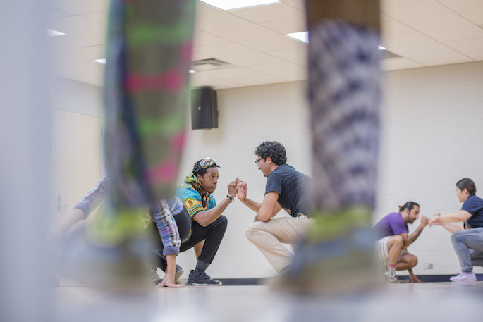 AMC attendees performing a duet activity crouched down and facing each other with one hand up touching the other, framed between a close-up of a person's legs