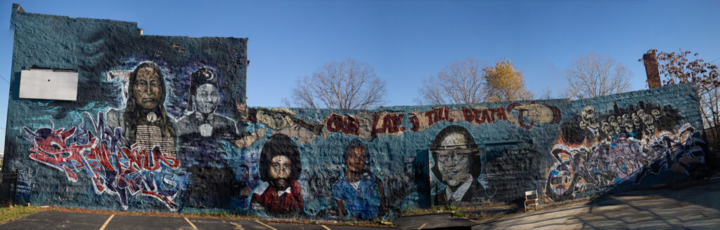 A panorama picture of the mural titled "Our Land Till Death Do Us Part" on the full side of a long building featuring images of  Malice Green, Aiyana Stanley-Jones, Vincent Chin, Crazy Horse, and Elijah Muhammad