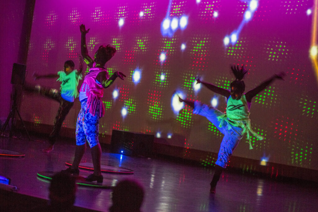 three dancing people in neon blacklit clothing with a constellation of glowing lights behind them.