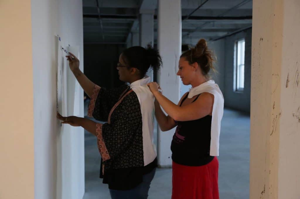 In a large dim naturally lit room with big white columns, two people stand in front of a white wall. Closest to the wall, a femme-presenting dark-skinned person draws on a sheet of paper. Behind them a light-skinned femme-presenting person mirrors their gesture and draws onto a piece of white fabric pinned to the first person’s back like a cape.