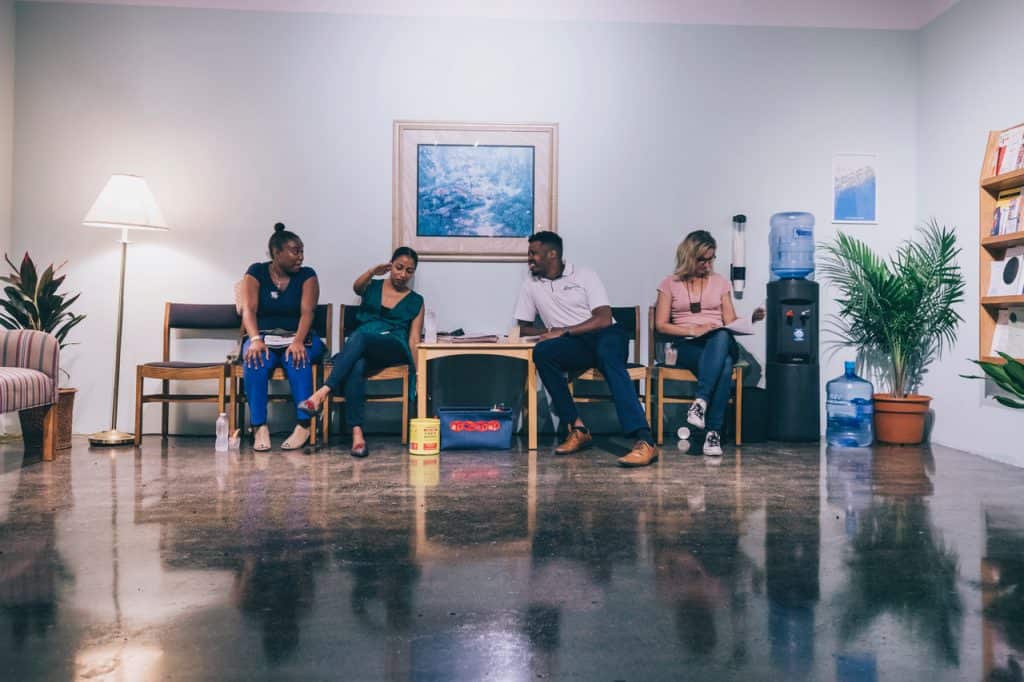 Four people sit in dark-cushioned waiting room chairs against a light-green wall. An illuminated standing lamp and a green plant are in the left corner corner. In the right corner is a water cooler and a potted palm next to a wall-mounted brown bookshelf full of pamphlets. A framed serene landscape and a blue poster hang on the wall.