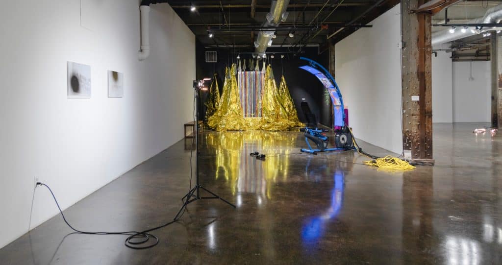 View onto a white-walled gallery space with grey polished concrete floors, exposed steel beams, and silver ceiling ventilation shafts. In the foreground is a flood light atop a three-legged light stand next to two photographs with black splotches scattered across them. In the distance is a black wall and tented metallic gold fabric hanging from the ceiling with a rainbow colored glittery curtain in front. In the center of the floor is a chroma-key blue exercise bike with four video monitors curving up a rod. 