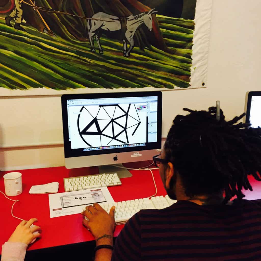 A Black man with dreads working on a computer, creating logo for their organization at the AMC near other people working on computers