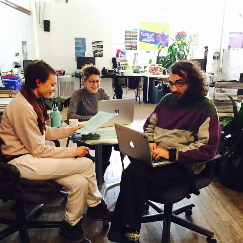 Three people working together sitting in roller chairs, two on laptops the other holding a piece of paper, all smiling