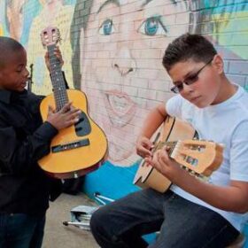 Two children playing guiter infront of a mural of the closeup of a child's face