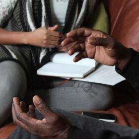 Closeup of hands gesturing, with another person sitting with a notebook on a table in the background