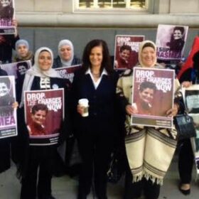 A woman standing holding coffee next to other women holding signs of a person and the words "Drop the charges now"