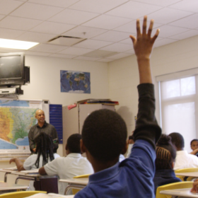 Image from behind a young Black student raising their hand with the teacher at the head of the class