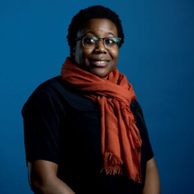 photo of Moya Bailey against a deep blue background, wearing a black shirt and brick red scarf