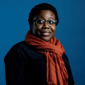photo of Moya Bailey against a deep blue background, wearing a black shirt and brick red scarf