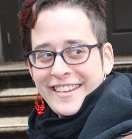 Headshot of Hannah Sassaman wearing red earrings and gray scarf