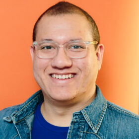 smiling light skinned Black and Asian man wearing a deep blue t-shirt under a denim button up on front of an orange background.