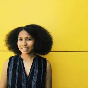 a black femme person with a v-neck shirt smiling in front of a yellow background