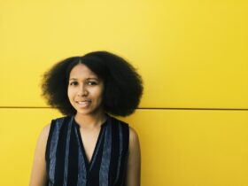 a black femme person with a v-neck shirt smiling in front of a yellow background