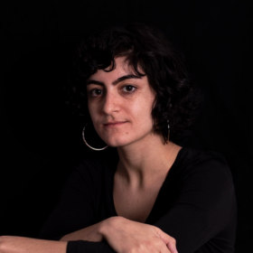 A palestinian femme person with chin length dark hair, wearing black in front of a black background