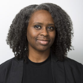 a black woman with shoulder-length hair wearing a black shirt and blazer in front of a grey background0