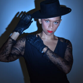 a femme person wearing dark gloves, a lace shirt, dark tilted hat, and red lipstick stands before a blue background