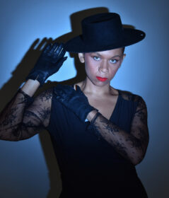 a femme person wearing dark gloves, a lace shirt, dark tilted hat, and red lipstick stands before a blue background