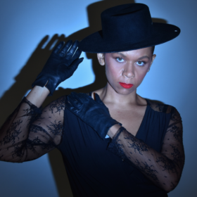a femme person wearing dark gloves, a lace shirt, dark tilted hat, and red lipstick stands before a blue background