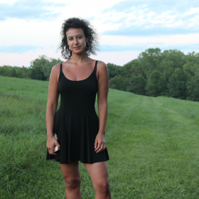 A brown-skinned woman stands toward the left of the frame wearing a fitted black sleeveless dress that cuts mid-way across her thighs. She has black curly hair and looks at the camera with a slight smirk. Behind her is a landscape with a sloping hill covered in lush green grass and a bright blue sky dotted with fluffy white clouds.