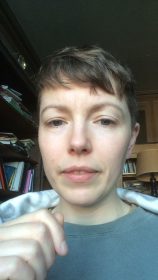 A selfie of a pale white person in winter light sitting at a desk wrapped in a blanket.